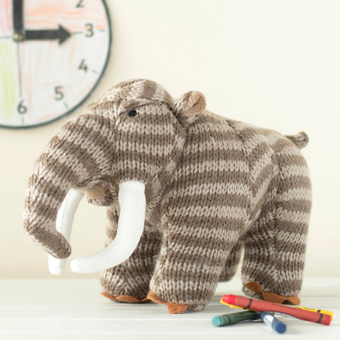 Knit plush woolly mammoth with alternating light and dark brown stripes.  It is on a table with some crayons and a clock on the wall behind it.