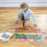 Young child in khaki pants and long sleeve blue shirt with dinosaurs on it playing with the puzzle on a hardwood floor.
