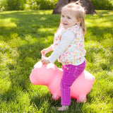 Young child bouncing on the Pink Pig Farm Hopper in a yard of bright green grass dappled by a large shade tree in the background.