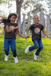 Two young children jumping down a grassy hill wearing white sneakers, blue jeans, and the Allosaurus tee shirt.