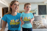 Mother with her arm around her toddlers waist.  Both are intently looking at something out of view.  They are in a living room with couch and view of kitchen area in the background.  Both are wearing Scooby-Doo! Mansion Mayhem tee shirts.  